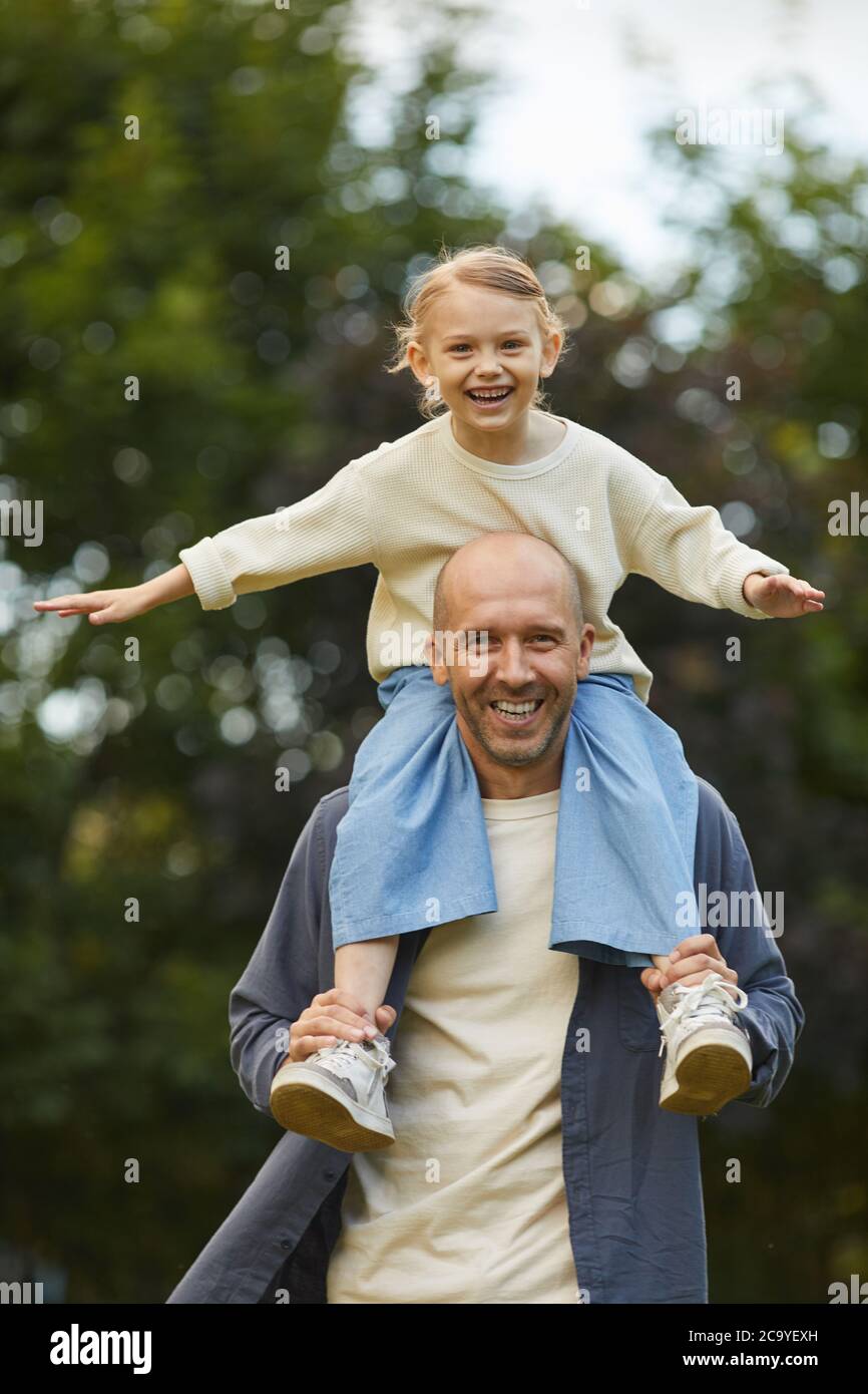 Portrait vertical d'une jolie fille assise sur les épaules des papas et s'amusant tout en appréciant la marche dans le parc à l'extérieur, les deux souriant à l'appareil photo Banque D'Images