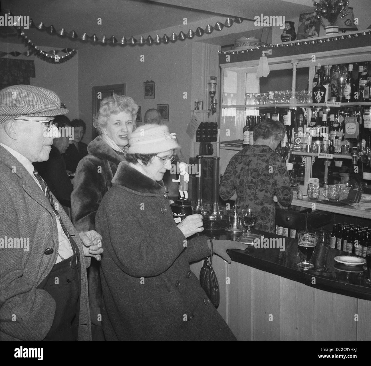 Années 1950, historique, deux dames en manteau, portant un manteau de  fourrure, ayant une boisson, un verre de sherry, dans un bar d'un pub, avec  un homme debout portant une casquette en