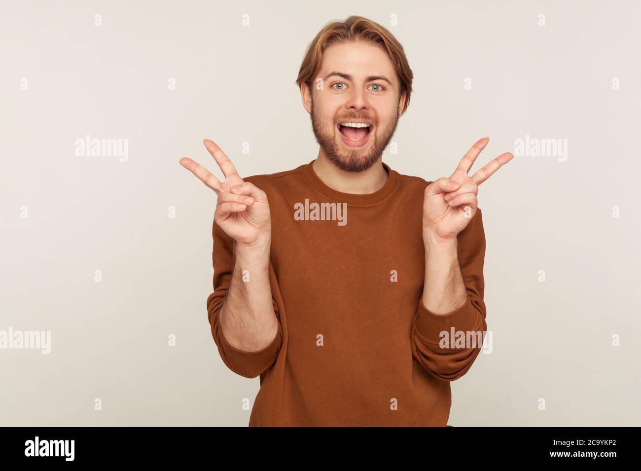 Portrait de l'homme heureux optimiste réussi barbu en sweat-shirt montrant le geste du doigt de victoire et criant pour la joie, célébrant la victoire, faisant le double Banque D'Images