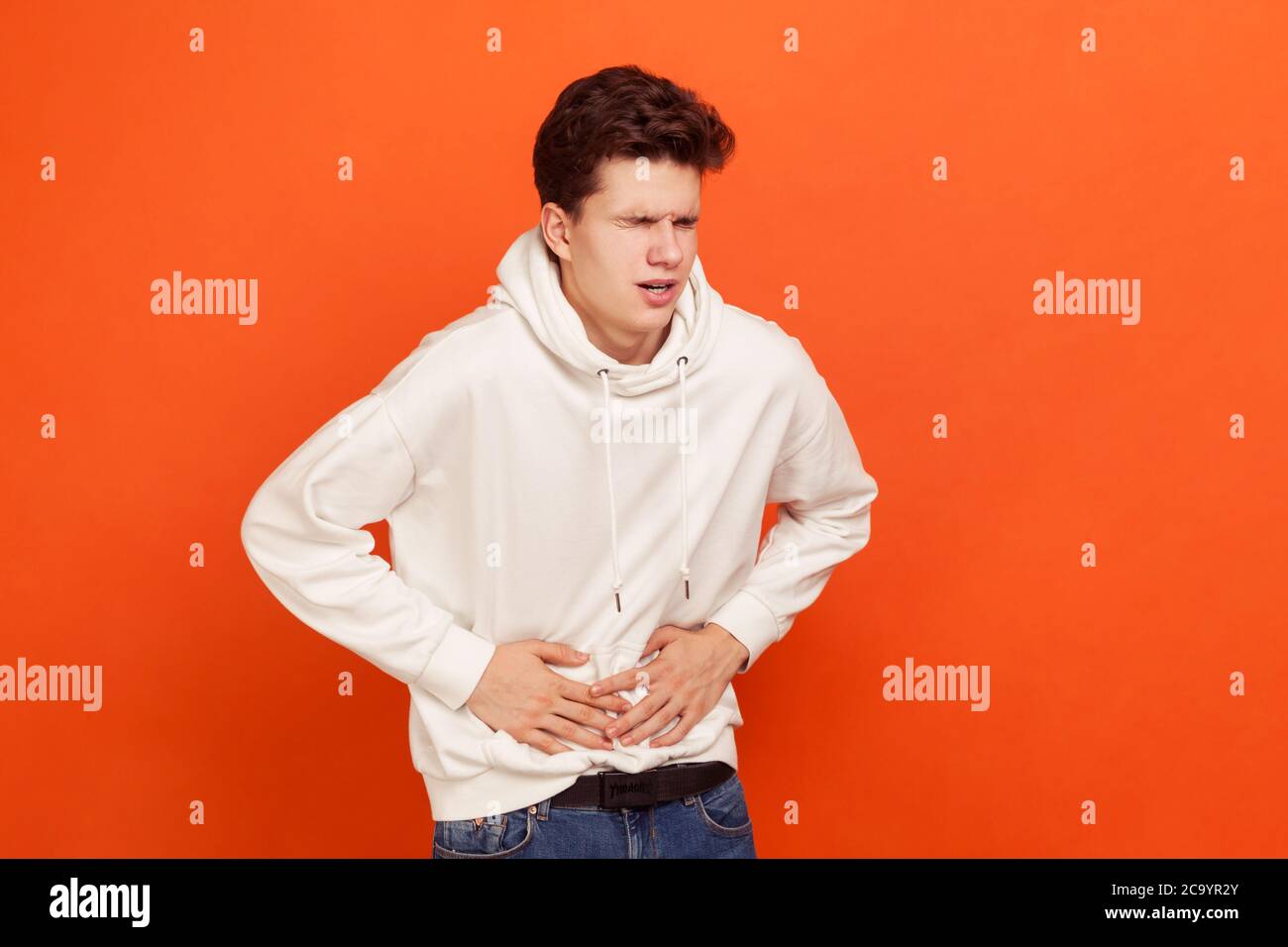 Nerveux jeune homme tendu dans un sweat-shirt décontracté avec capuche tenant les mains sur l'estomac, sensation de terrible douleur abdominale et spasmes, diarrhée, nausée. Intérieur Banque D'Images