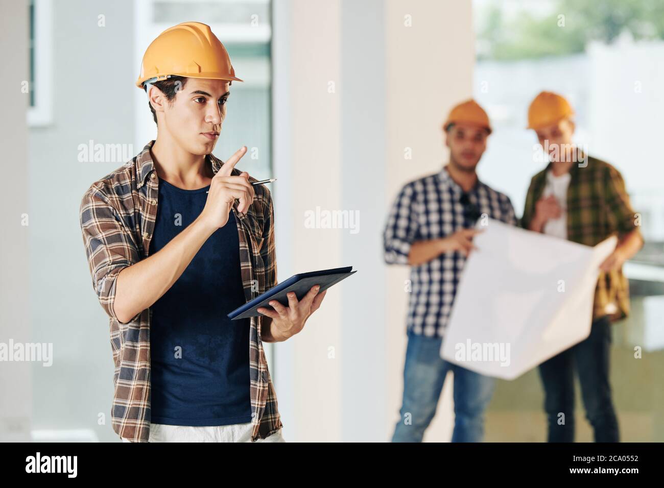 Beau jeune ingénieur civil en casque et chemise à carreaux inspectant le site de construction et prenant des notes dans le document sur l'ordinateur tablette Banque D'Images