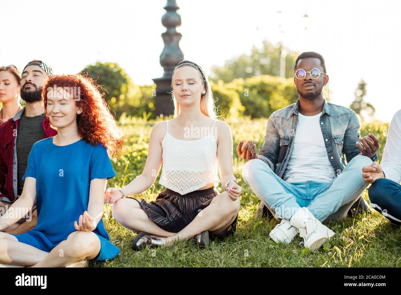 Divers cours de yoga de jeunes amis gaies en été vêtements décontractés assis dans lotus poser sur vert lown dans le parc public, vue de face avec l'accent sur fe Banque D'Images