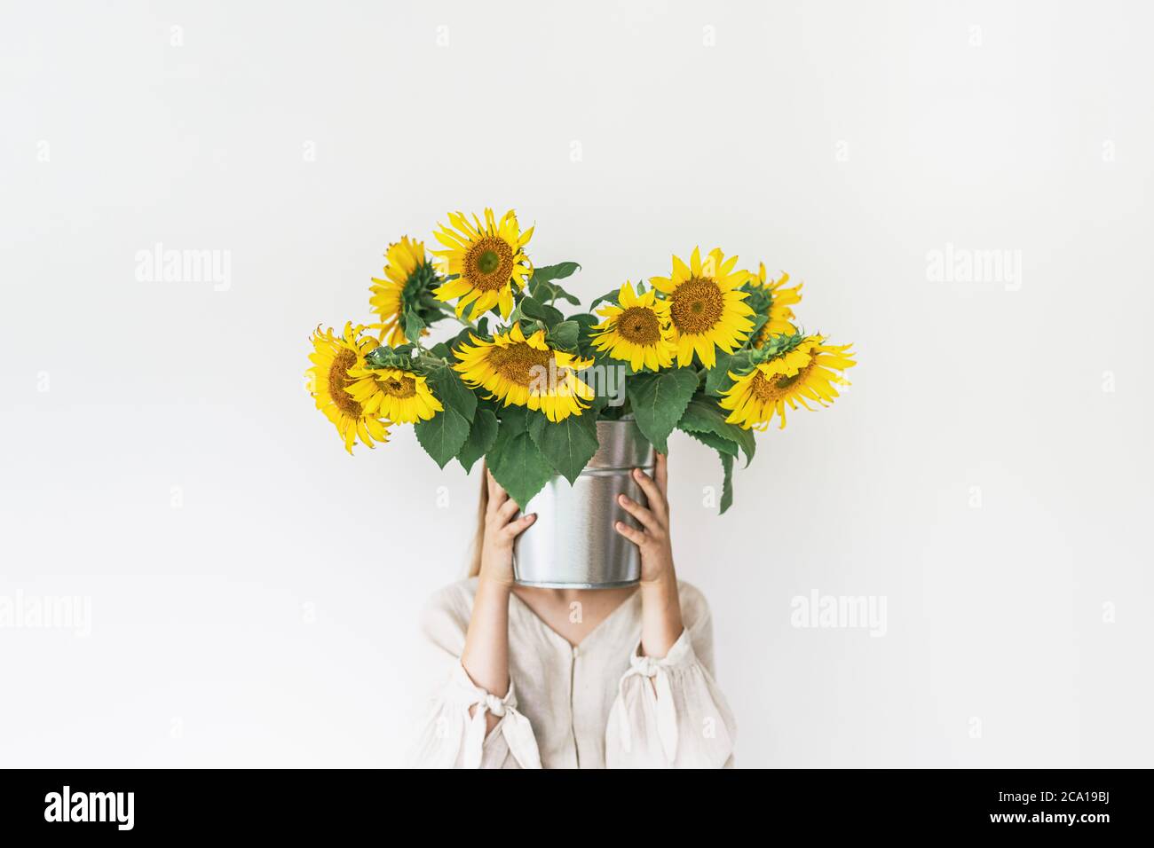 Belle jeune femme en robe de lin tient le bouquet de tournesol dans un seau en étain sur fond blanc. Banque D'Images