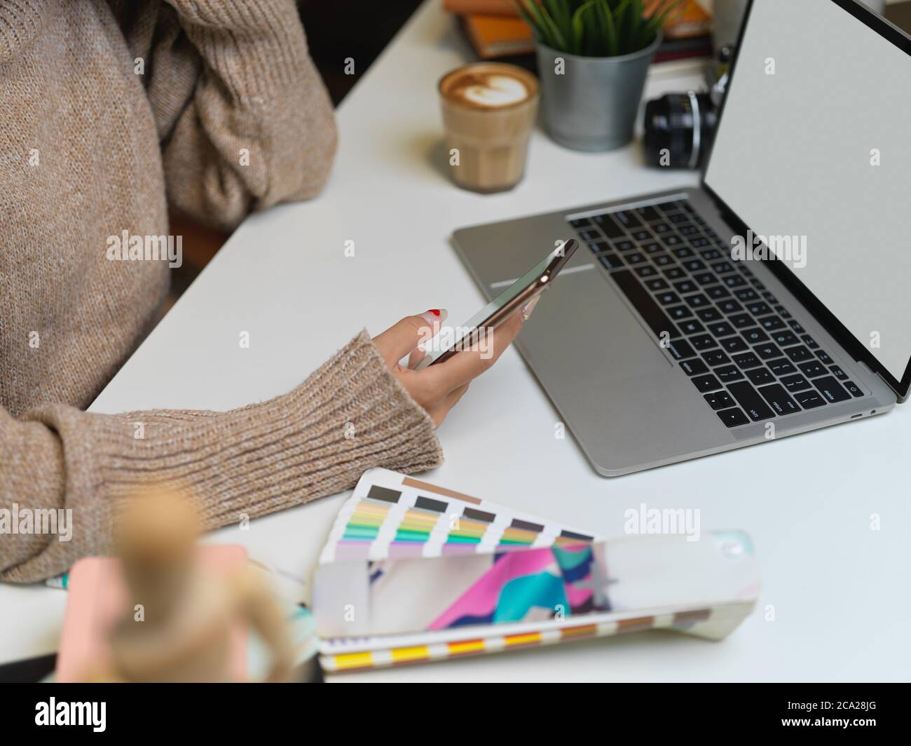 Photo courte d'une femme de créateur dans un chandail à l'aide d'un smartphone tout en travaillant avec un ordinateur portable factice sur le bureau Banque D'Images