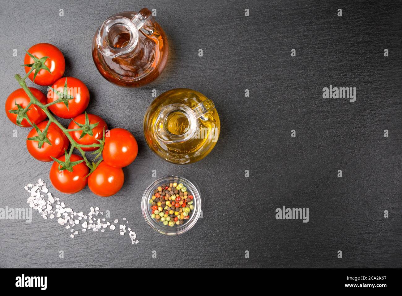 Une brindille de tomates cerises avec quelques grains de sel épais, une huile et une carafe de vinaigre et du poivre coloré sur une dalle d'ardoise. Avec espace de copie. Banque D'Images