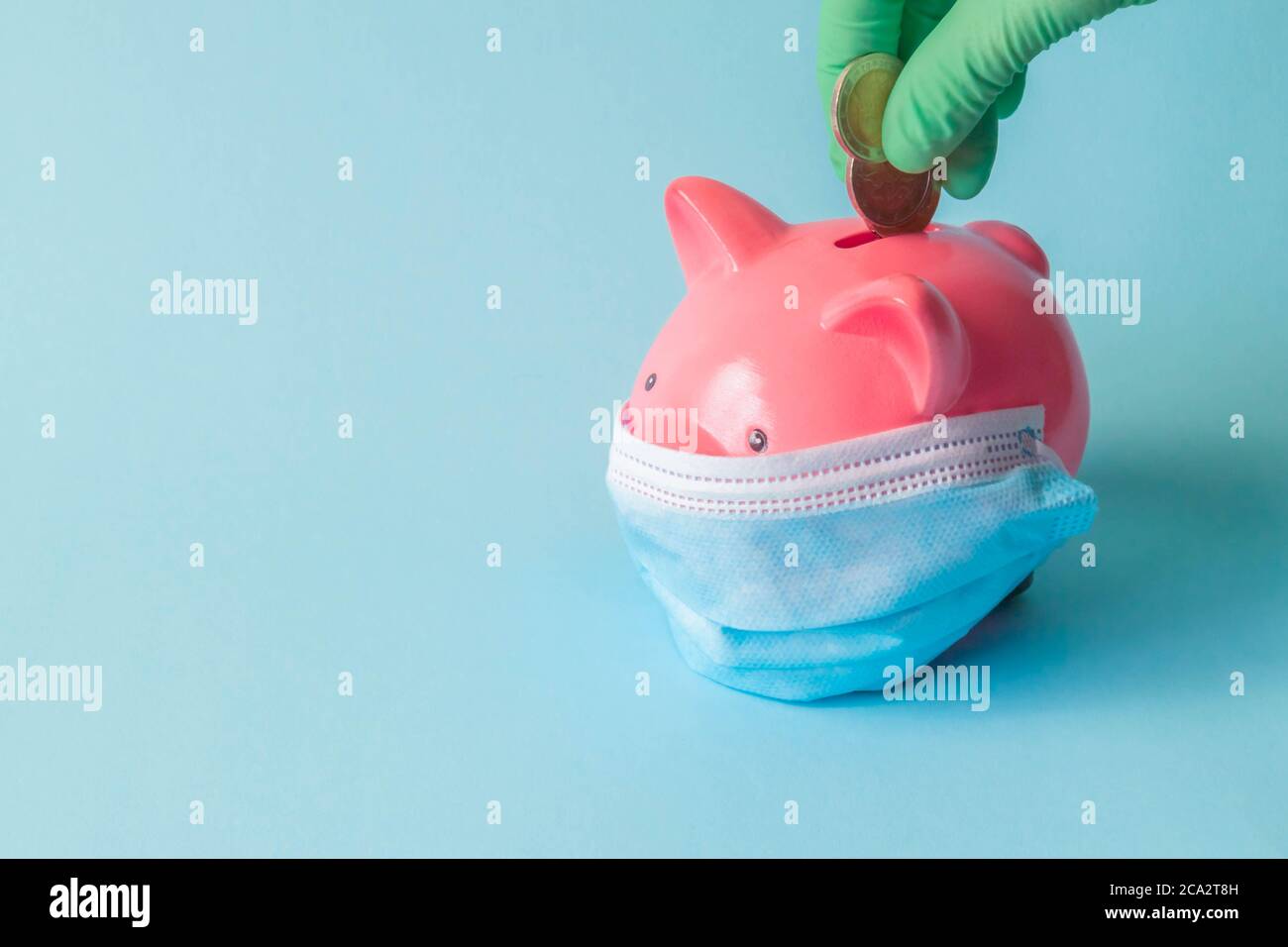 Tirelire avec masque protecteur et main dans le gant mettant des pièces en euros sur un fond isolé. La main dans le gant pute l'argent dans la banque de porc avec le masque. Banque D'Images