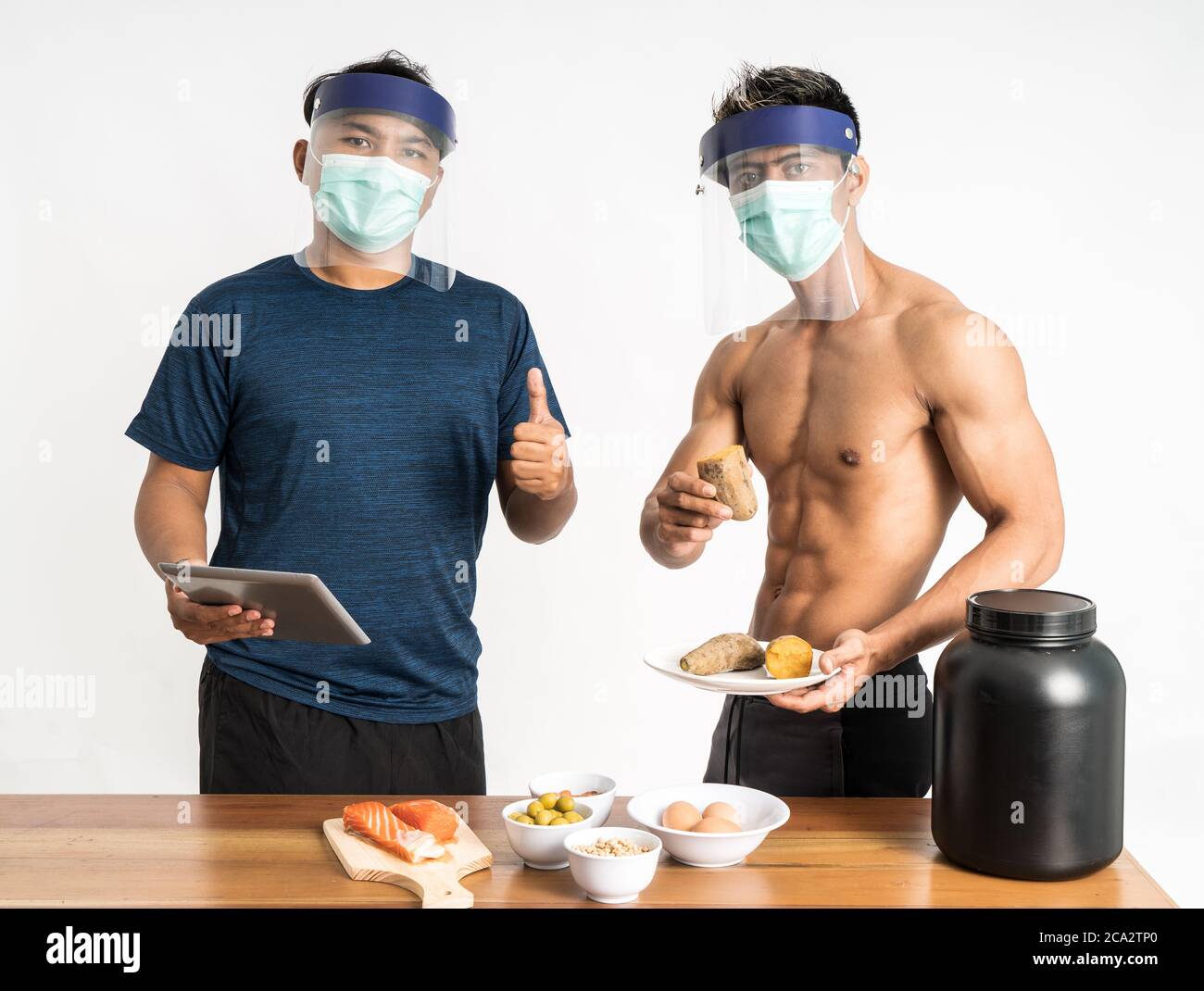 deux blogueurs asiatiques de santé et de sport portant un masque et écran facial avec les pouces vers le haut montrant le menu des aliments sains pendant tenez un support de tablette orienté vers l'avant et regardez l'appareil photo sur un arrière-plan isolé Banque D'Images