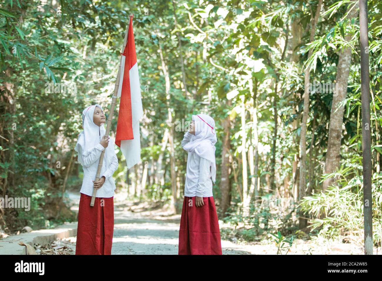 Deux petites filles musulmanes portant des voiles portant un drapeau rouge et blanc et l'autre fille se saluant sur un fond d'arbre Banque D'Images