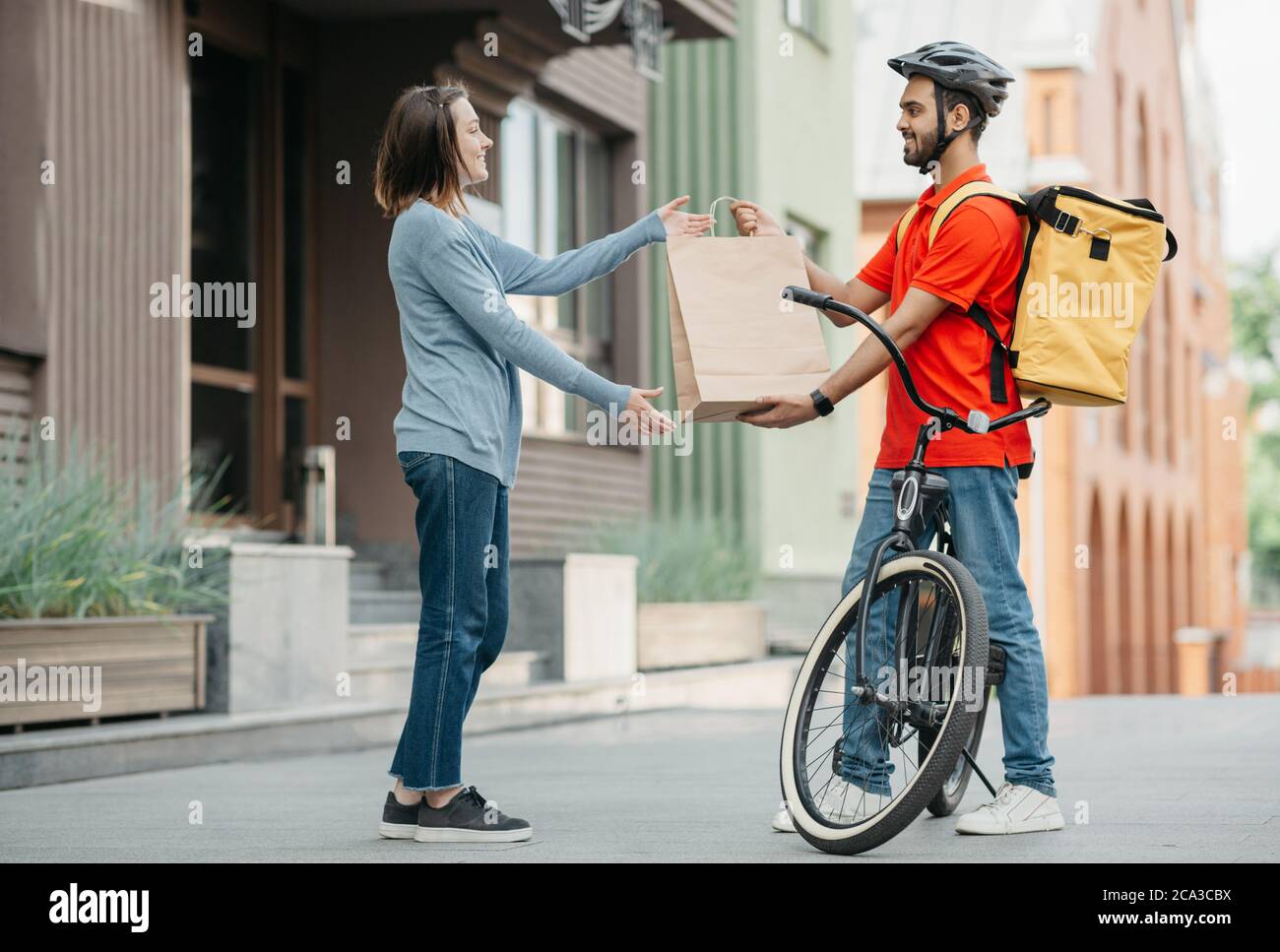 Un transporteur avec vélo a livré le sac à provisions au client. Une fille souriante prend le sac de l'homme dans un casque avec sac à dos Banque D'Images