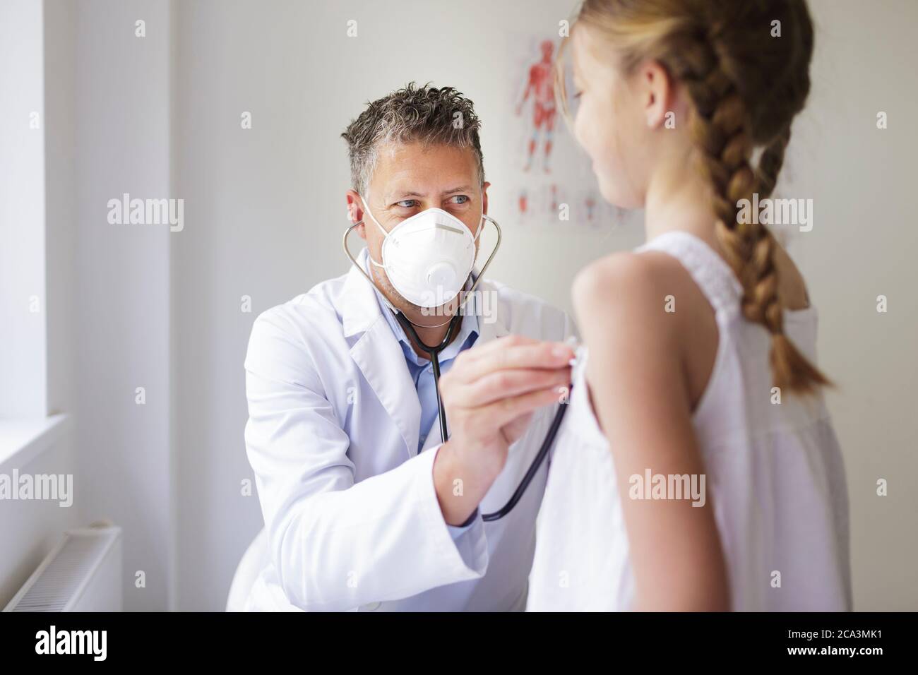 un bon médecin de sexe masculin avec une barbe de trois jours et un masque de nez et de bouche examine une jeune fille avec un stéthoscope Banque D'Images