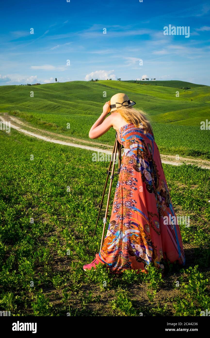 Vallée d'Orcia, quartier de Sienne, Toscane, Italie, Europe. Femme blonde avec chapeau prend des photos dans les champs verts. Banque D'Images