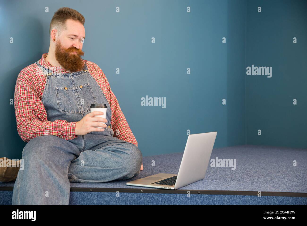 Portrait d'un boxeur à barbe brutale dans une combinaison bleue, une chemise à carreaux, boire un café dans une tasse en papier travaillant sur un ordinateur portable assis près du mur. Dist Banque D'Images