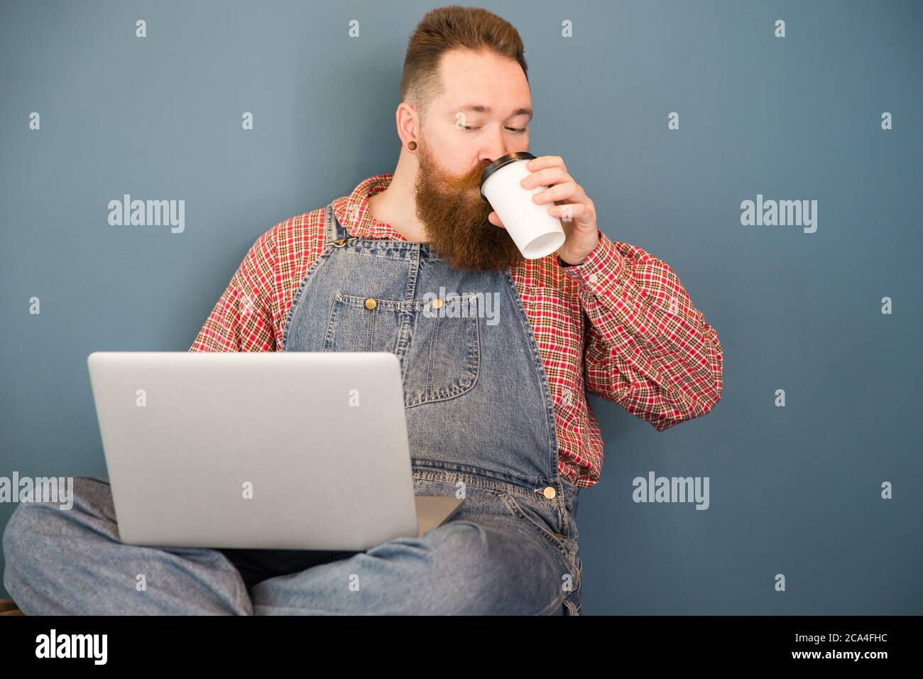 Portrait d'un boxeur à barbe brutale dans une combinaison bleue, une chemise à carreaux, boire un café dans une tasse en papier travaillant sur un ordinateur portable assis près du mur. Dist Banque D'Images