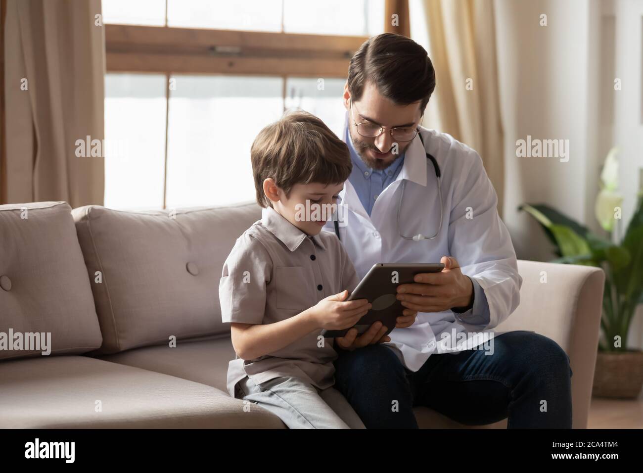 Jeune pédiatre sympathique montrant des dessins animés sur une tablette à un petit patient. Banque D'Images