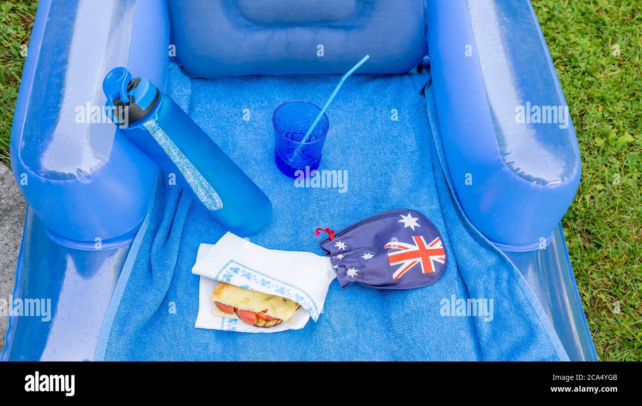 Composition dans les tons bleus d'un pique-nique sur l'herbe avec matelas gonflable, bouteille d'eau, sandwich, verre avec paille et drapeau australien Banque D'Images