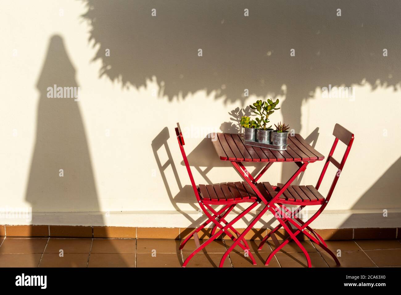 Table et chaises en bois rouge et en métal dans le jardin, à côté d'un mur blanc où vous pouvez voir les ombres d'un arbre et d'un parasol Banque D'Images