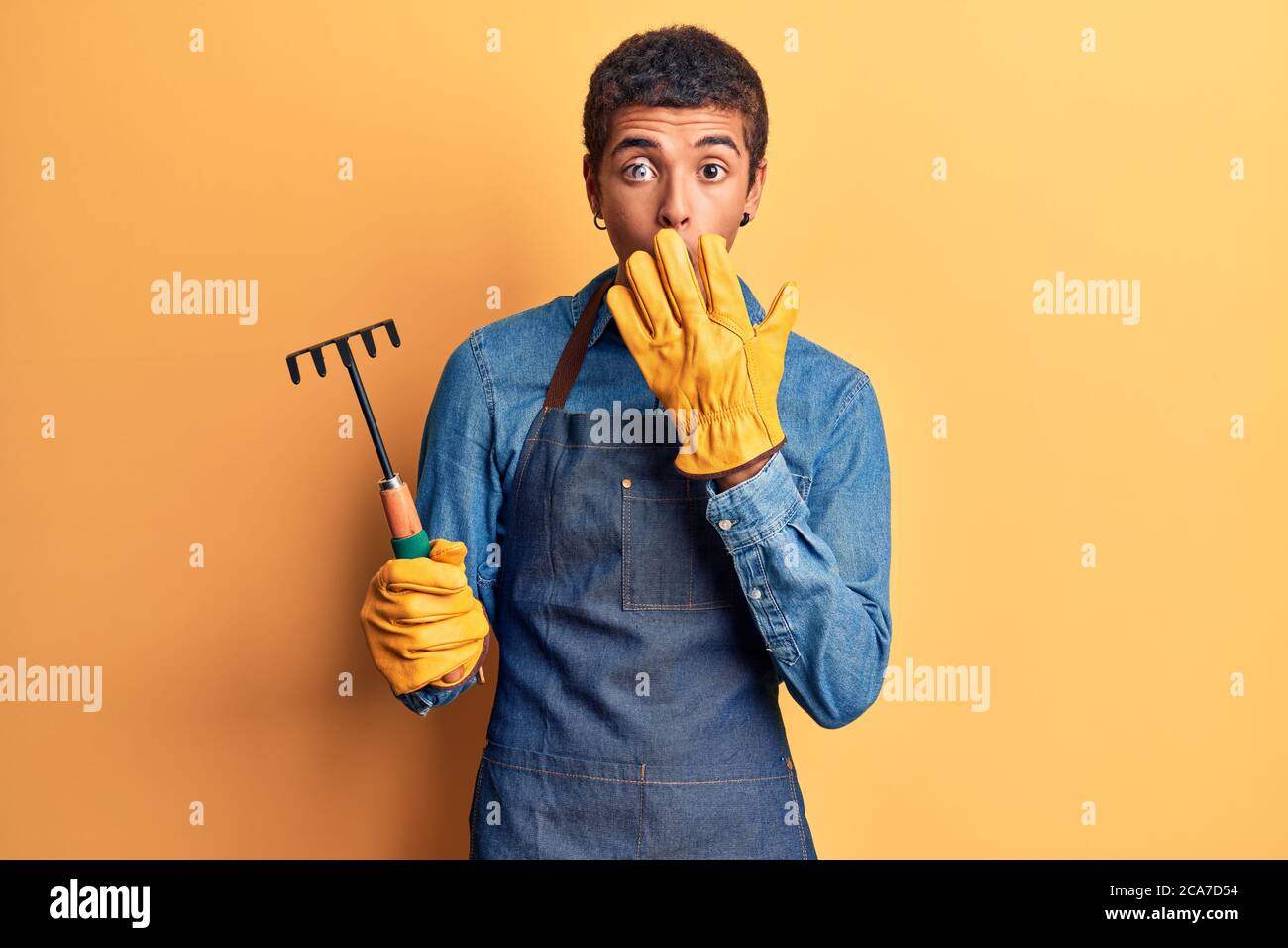 Jeune homme amérindien africain portant un tablier de jardinier et des gants tenant le râteau couvrant la bouche avec la main, choqué et peur d'erreur. Expressi surpris Banque D'Images