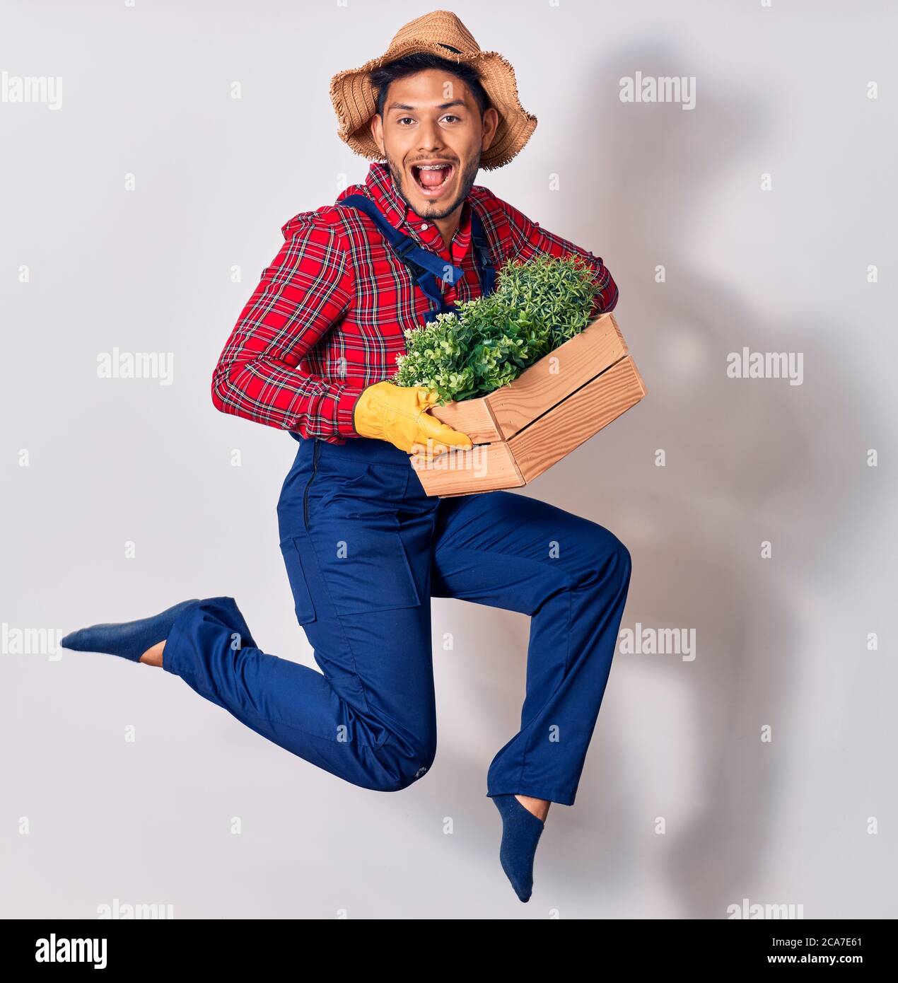 Jeune latins beau portant l'uniforme de fermier et chapeau souriant heureux. Sautant avec le sourire sur le visage plante pot en bois sur fond blanc isolé Banque D'Images