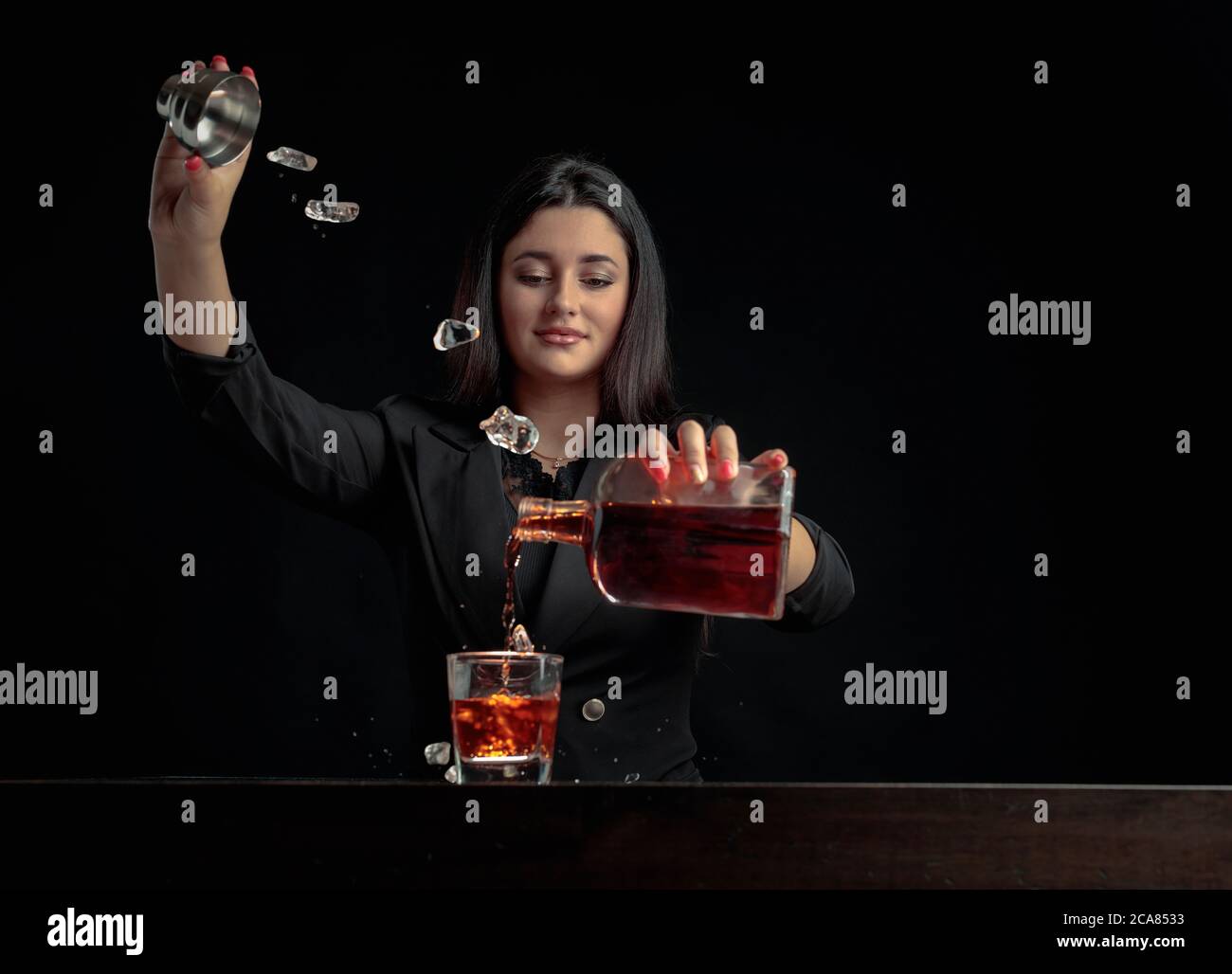 Une belle barman verse une boisson alcoolisée dans un verre et jette des morceaux de glace. Magnifique brunette en noir préparer un cocktail. Copier l'espace. Banque D'Images