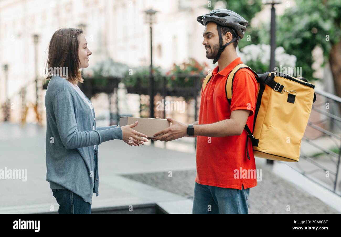Femme prend l'achat en ligne de messagerie. Livreur dans casque, avec sac à dos donne boîte au client Banque D'Images Femme prend l'achat en ligne de messagerie. Livreur dans casque, avec sac à dos donne boîte au client Banque D'Images