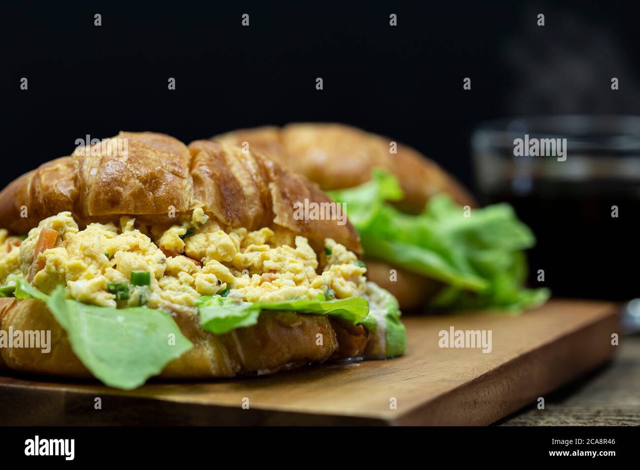 Gros plan sur des œufs brouillés farcis croissant sur un plateau en bois avec un fond de café chaud. Banque D'Images