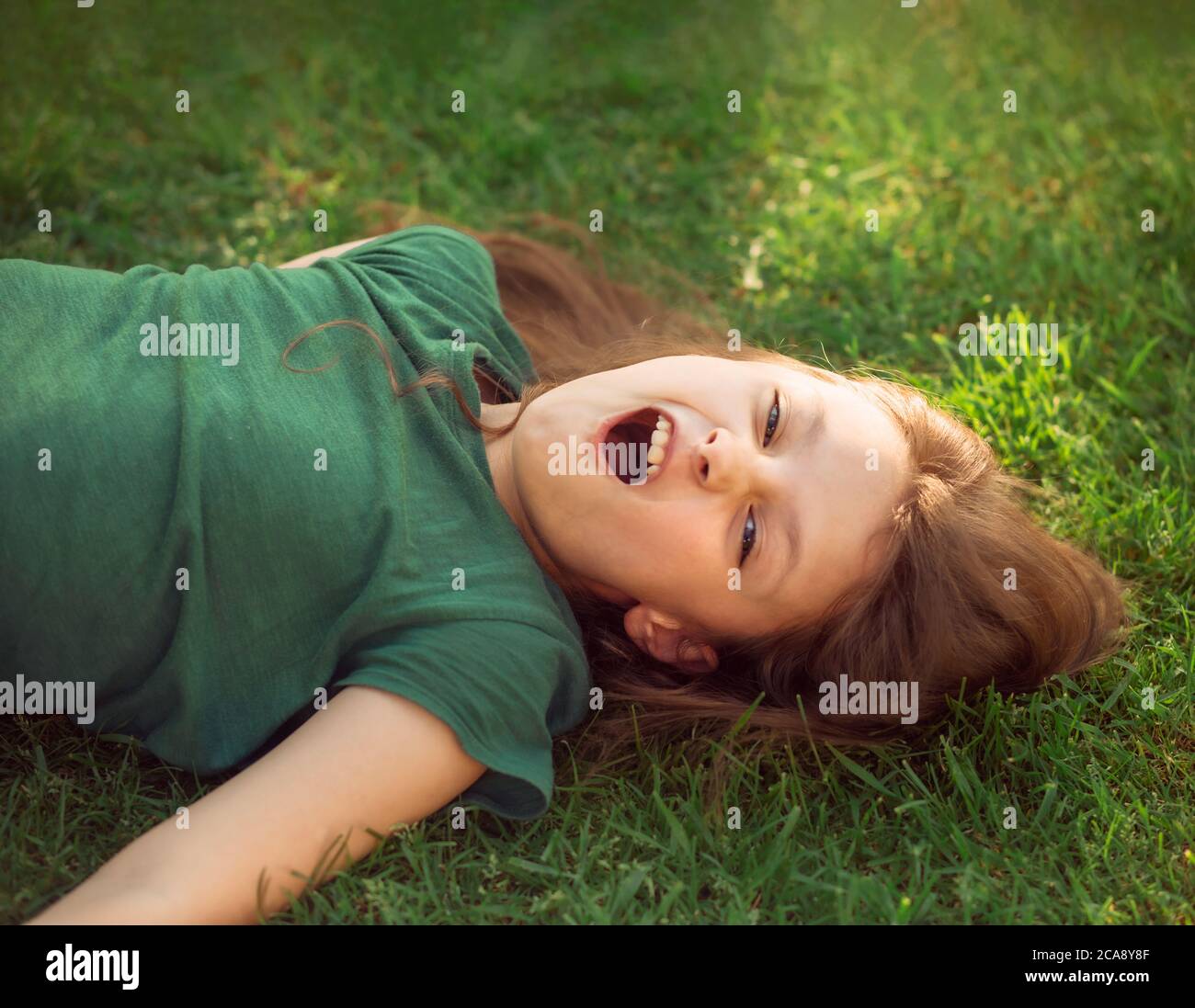 Riant amusant fou pleurs gamin fille couché sur l'herbe sur la nature fond d'été. Gros plan positif extérieur lumineux portrait ensoleillé Banque D'Images