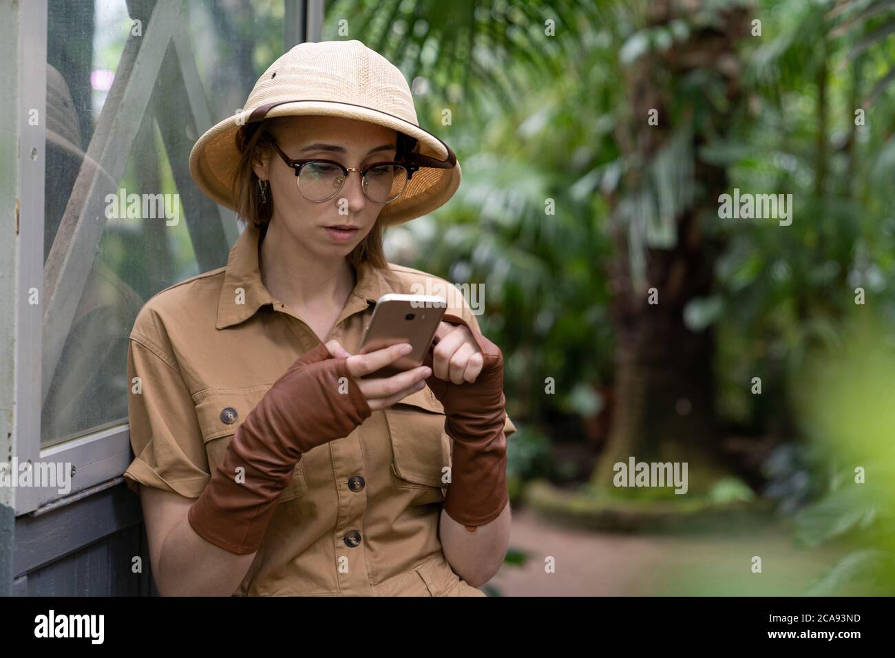 Femme botaniste vêtue de style safari en serre. Naturaliste en vêtements kaki, gants de castor pour se reposer et lire un livre, à l'intérieur. Banque D'Images