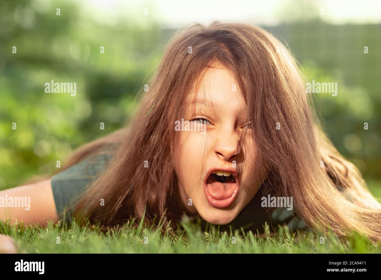 Une jeune fille folle en colère criant avec une bouche ouverte et des cheveux fous couchés sur l'herbe verte sur un fond d'été extérieur. Gros plan portrait émotionnel de diff Banque D'Images