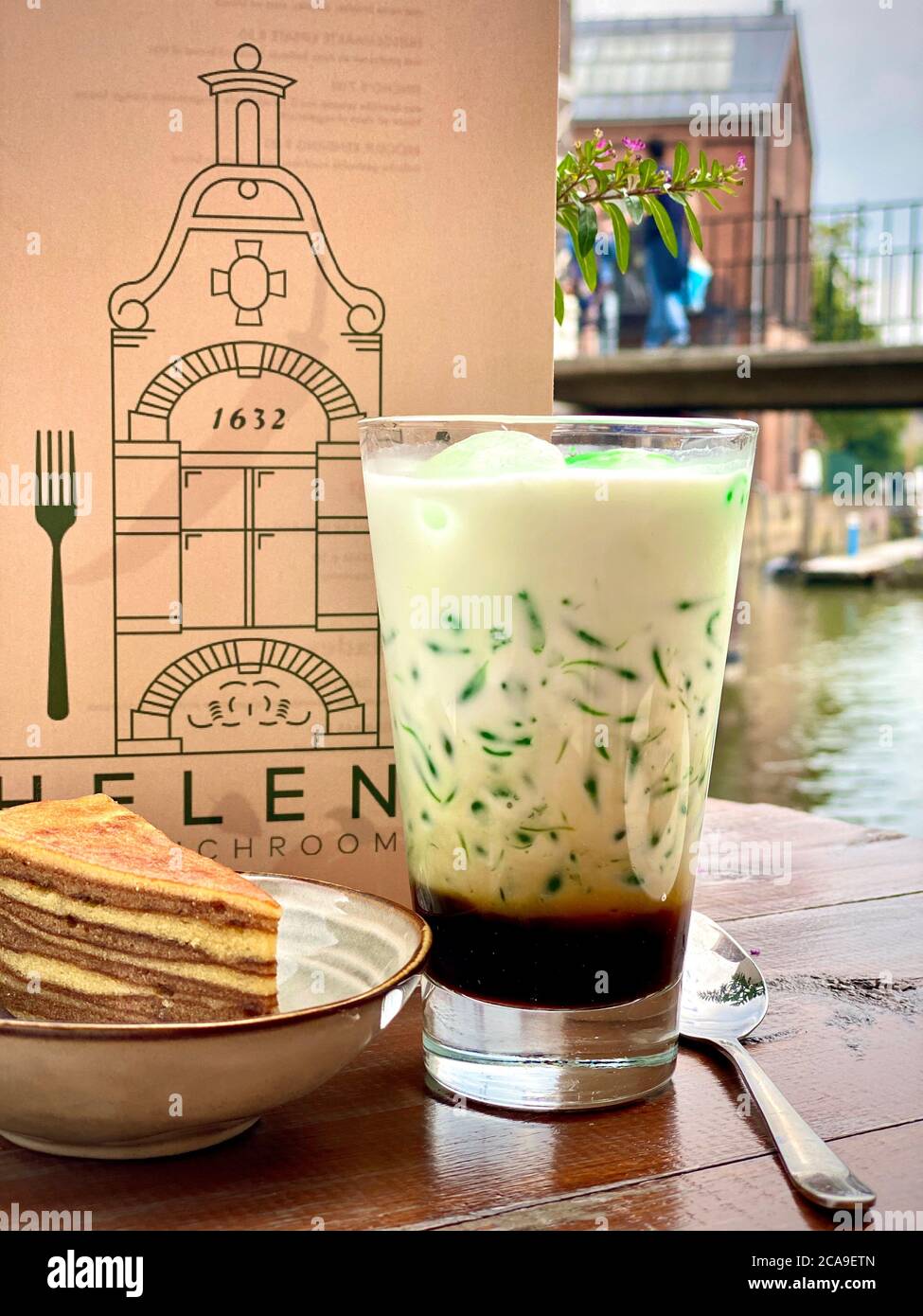 Cadol traditionnel indonésien à base de jus de pandas et de haricots mung. Restaurant café table en bois à l'extérieur. Boisson servie dans un verre avec une tranche de gâteau dans un bol avec un menu en arrière-plan. Boisson crémeuse colorée verte et brune. Soif assoiffée. Banque D'Images