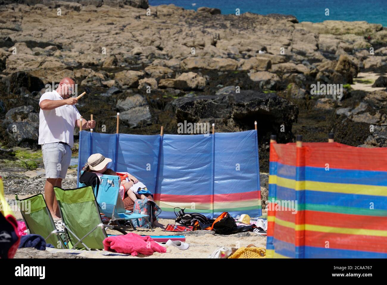 Homme érigeant une pause-vent sur la plage. Debout avec un maillet sur les poteaux Blue Windbreak femme assise sur la chaise de plage Boulders en arrière-plan. Format paysage Banque D'Images
