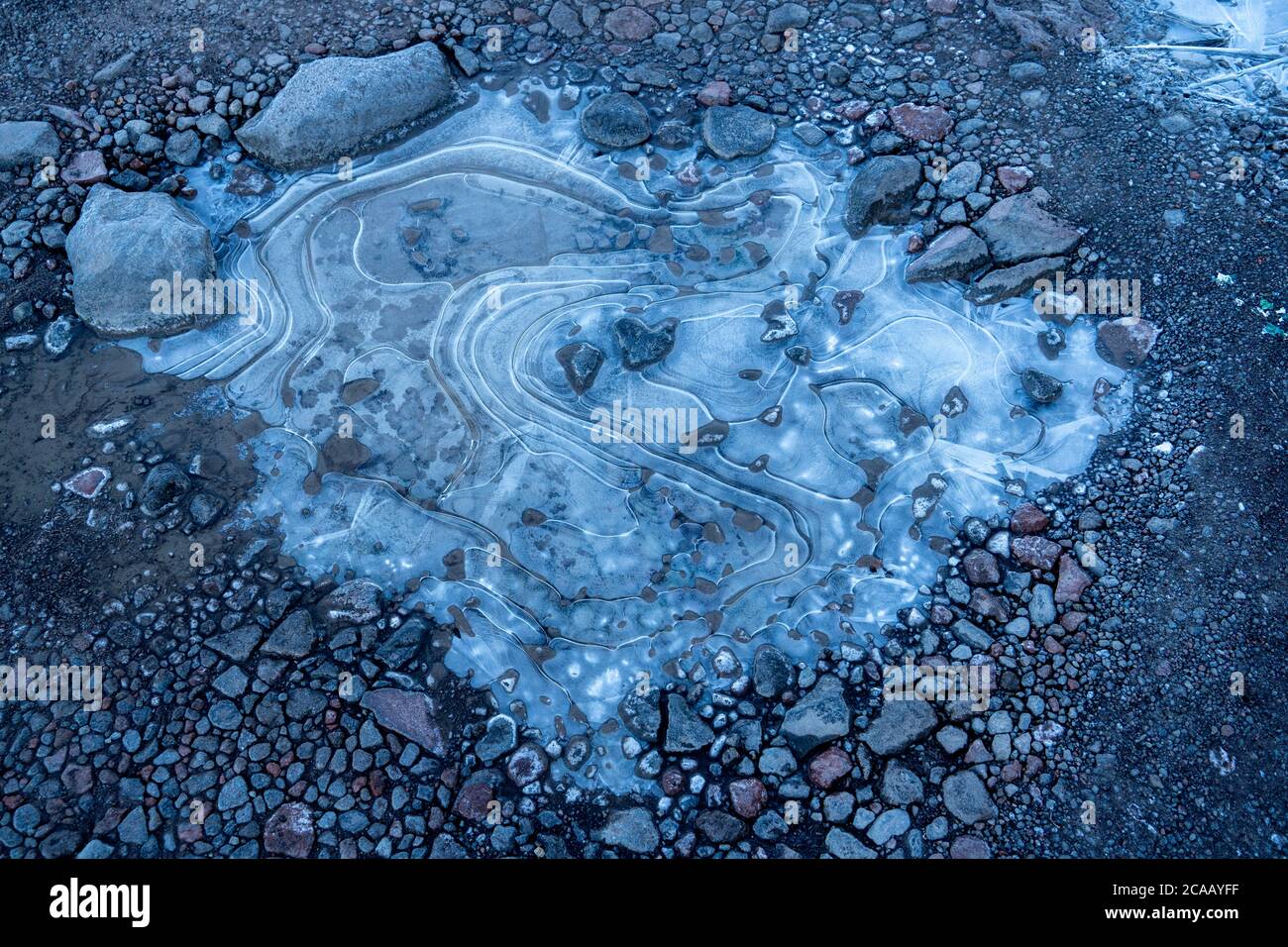 De l'eau dans une flaque gelée à des formes tourbillonnantes dans la glace. Fond de gravier et de cailloux. Banque D'Images