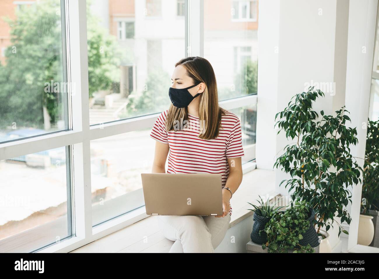 Jeune fille travailleur ou étudiant assis à la fenêtre avec un masque facial avec un ordinateur. Elle est restée à la maison ou travaille près de la fenêtre dans un espace ouvert. Banque D'Images