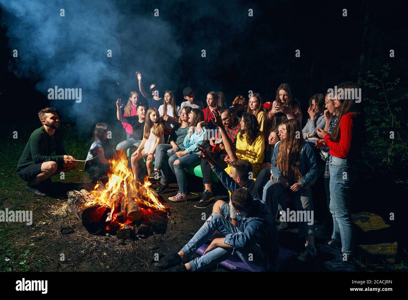 Vue latérale des enfants heureux assis sur un banc en bois et chantant près du feu de camp dans la chaude nuit d'été. Les Scoutmasters jouent de la guitare et s'amusent avec les enfants en forêt. Concept de camping d'été. Banque D'Images