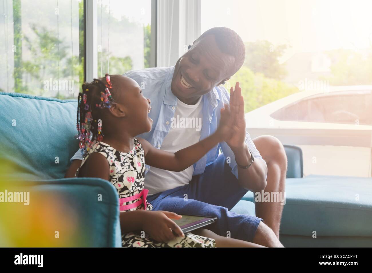 Père et fille africains utilisant une tablette à l'aspect heureux parents avec enfants se détendre à la maison dans un vêtement décontracté assis sur le canapé dans le salon Banque D'Images
