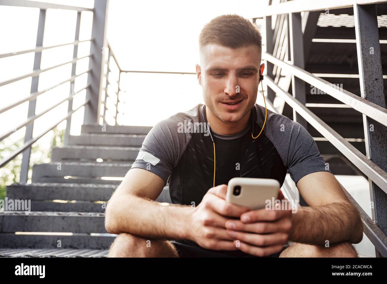 Image d'un sportif caucasien heureux utilisant un téléphone portable et des écouteurs tout en étant assis sur un escalier urbain à l'extérieur Banque D'Images