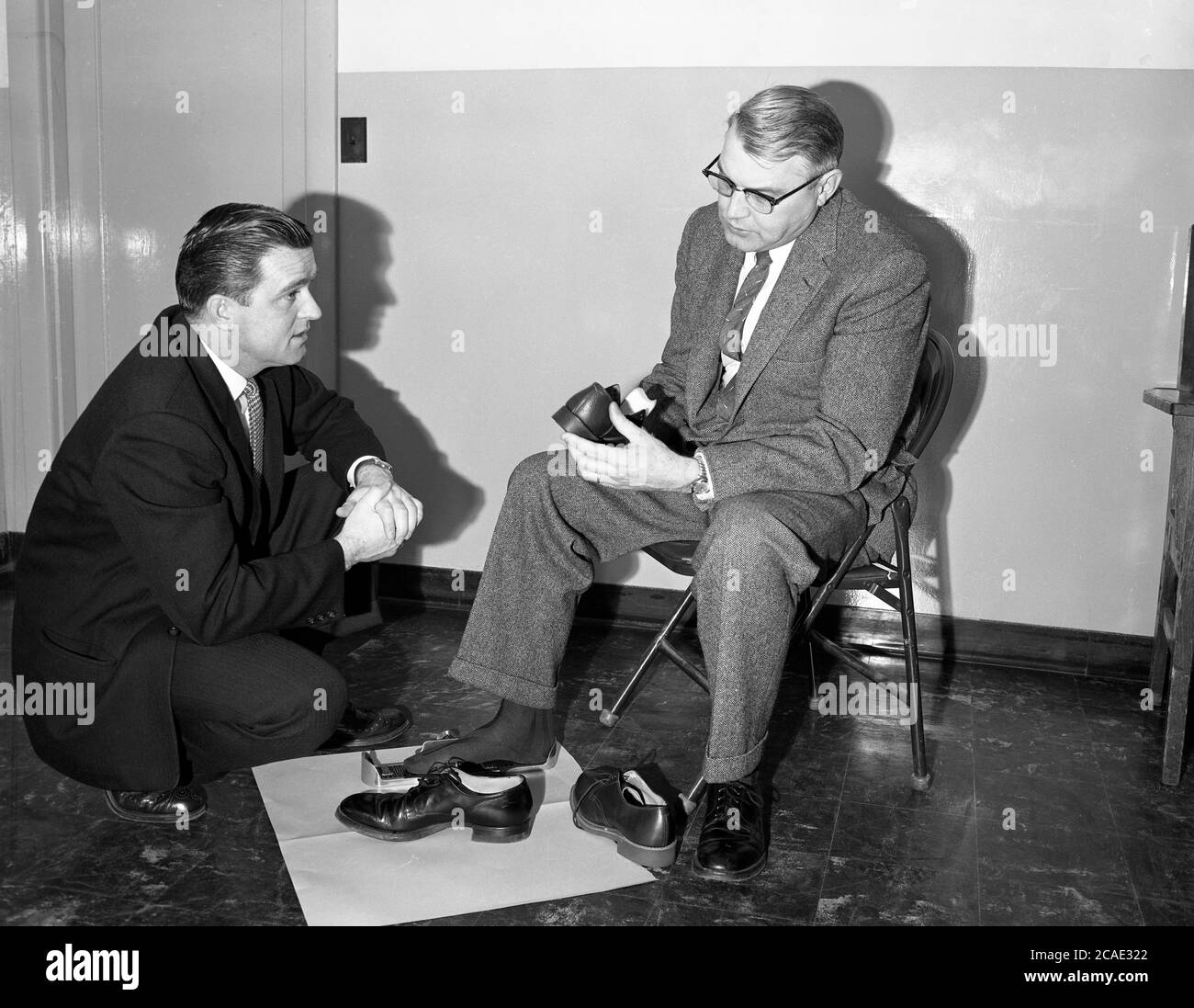 1940, historique, un vendeur de chaussures avec un client mâle dans une combinaison à carreaux intelligents considérant une nouvelle paire de chaussures. Le client a son pied droit dans une ancienne mesure de pied métallique ou une jauge de raccord, qui mesure la longueur et la largeur du pied. Connu sous le nom de Brannock Device, après son inventeur Charles F. Brannock, il a été introduit en 1927 et a été utilisé pour mesurer la taille de la chaussure d'une personne, en mesurant la longueur, la largeur et la longueur de la voûte plantaire du pied. Banque D'Images
