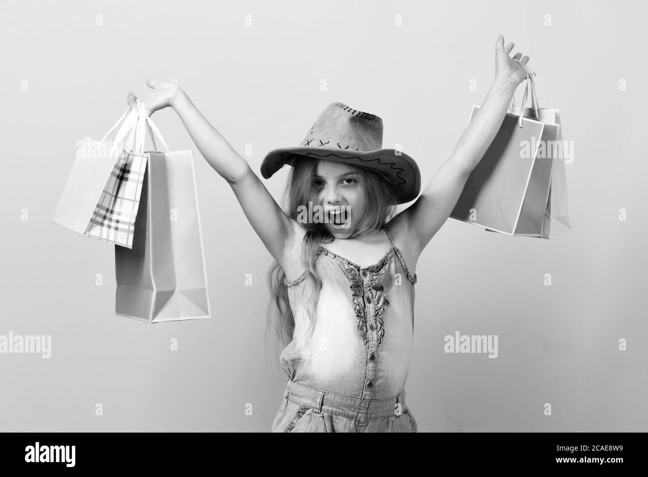 Enfant avec un visage confiant et heureux porte chapeau de cowboy et fait des achats. Fille avec des sacs de shopping roses. Concept de mode et de shopping. Fille d'école avec des paquets isolés sur fond rose Banque D'Images