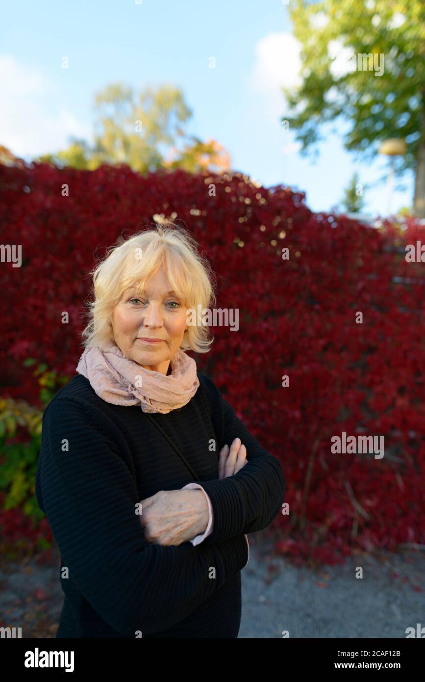 Femme âgée aux bras croisés devant des plantes rouges de super-réducteur couvrant la clôture Banque D'Images