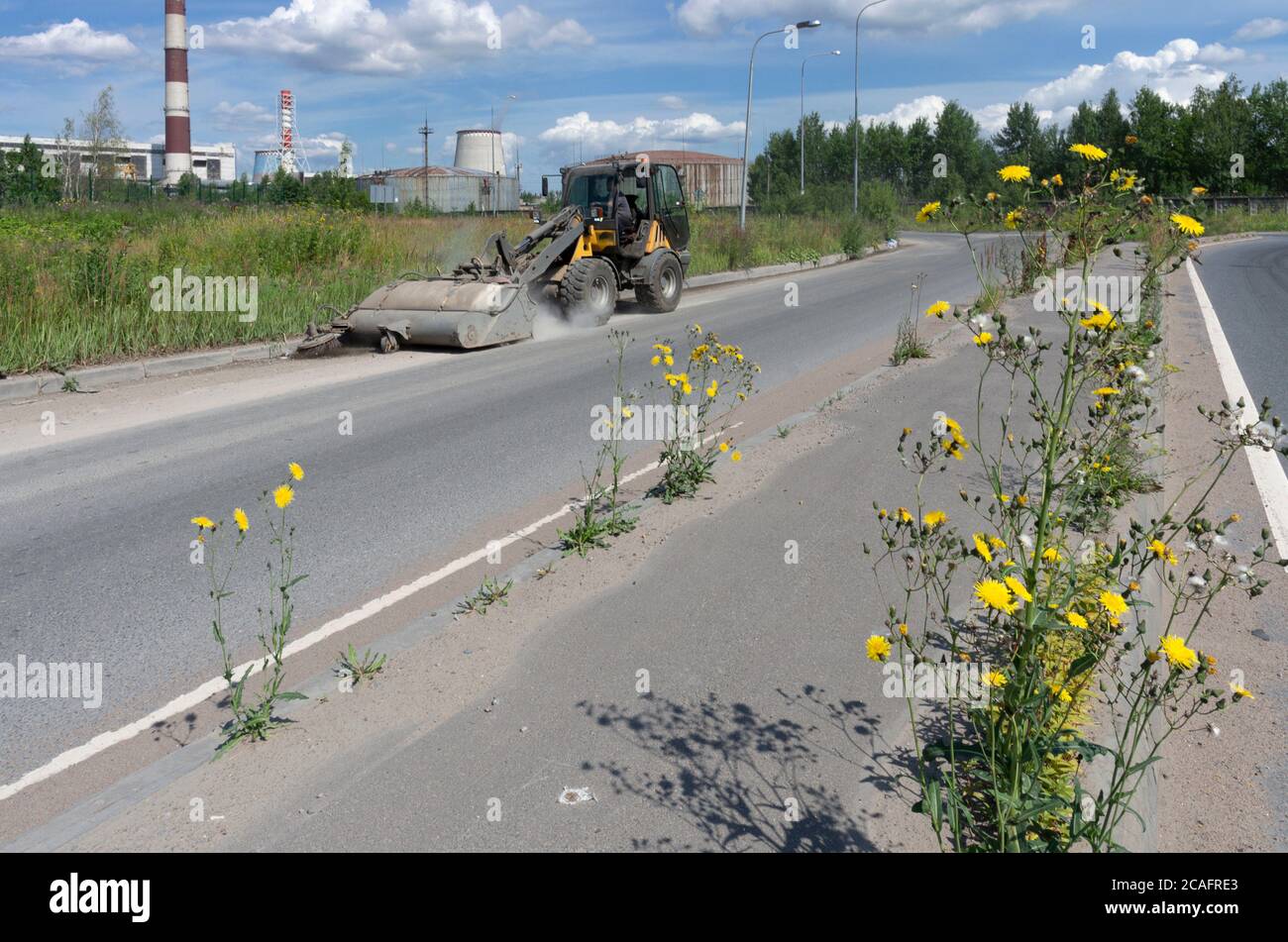 Le tracteur jaune élimine les déchets sur l'autoroute et les fleurs jaunes pousser sur le trottoir sur un fond industriel Banque D'Images
