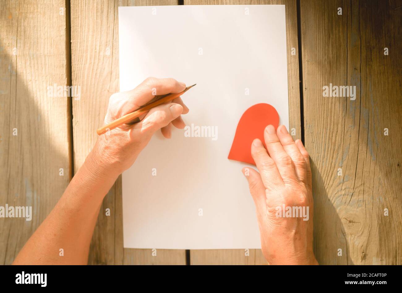 Femme âgée main allant écrire une lettre, coeur rouge sur table rustique en bois comme un fond, rayons du soleil. Vue de dessus, espace de copie. Mère ou femme, Banque D'Images