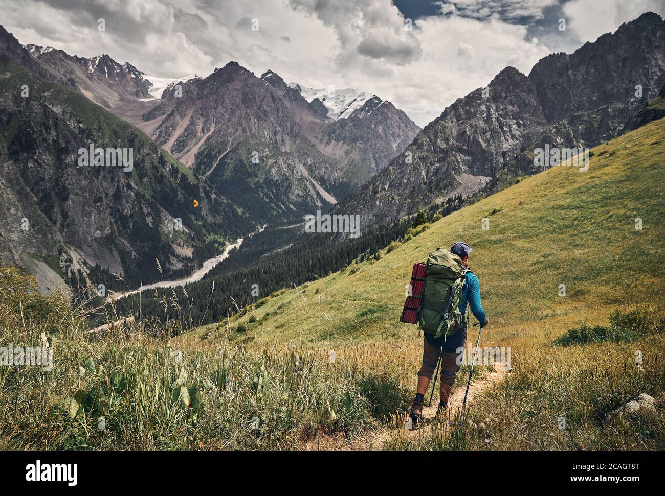Homme touristique avec grand sac à dos est de marcher dans la vallée de montagne verte avec des sommets enneigés et ciel nuageux fond Banque D'Images