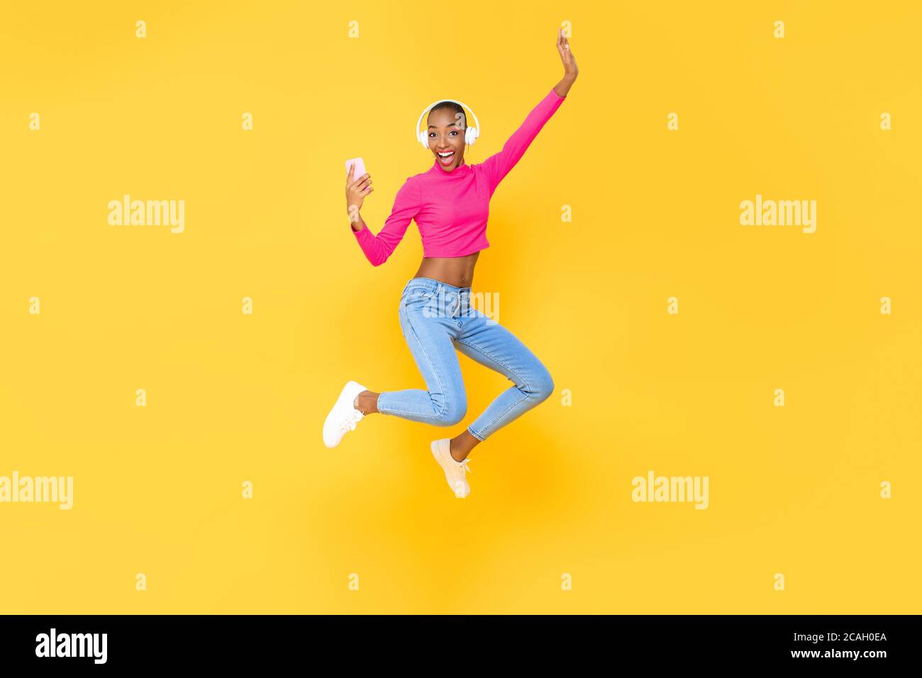 Femme afro-américaine pleine de sourire, énergique et vêtue d'un casque pour écouter de la musique du smartphone et du saut isolé sur fond jaune coloré Banque D'Images