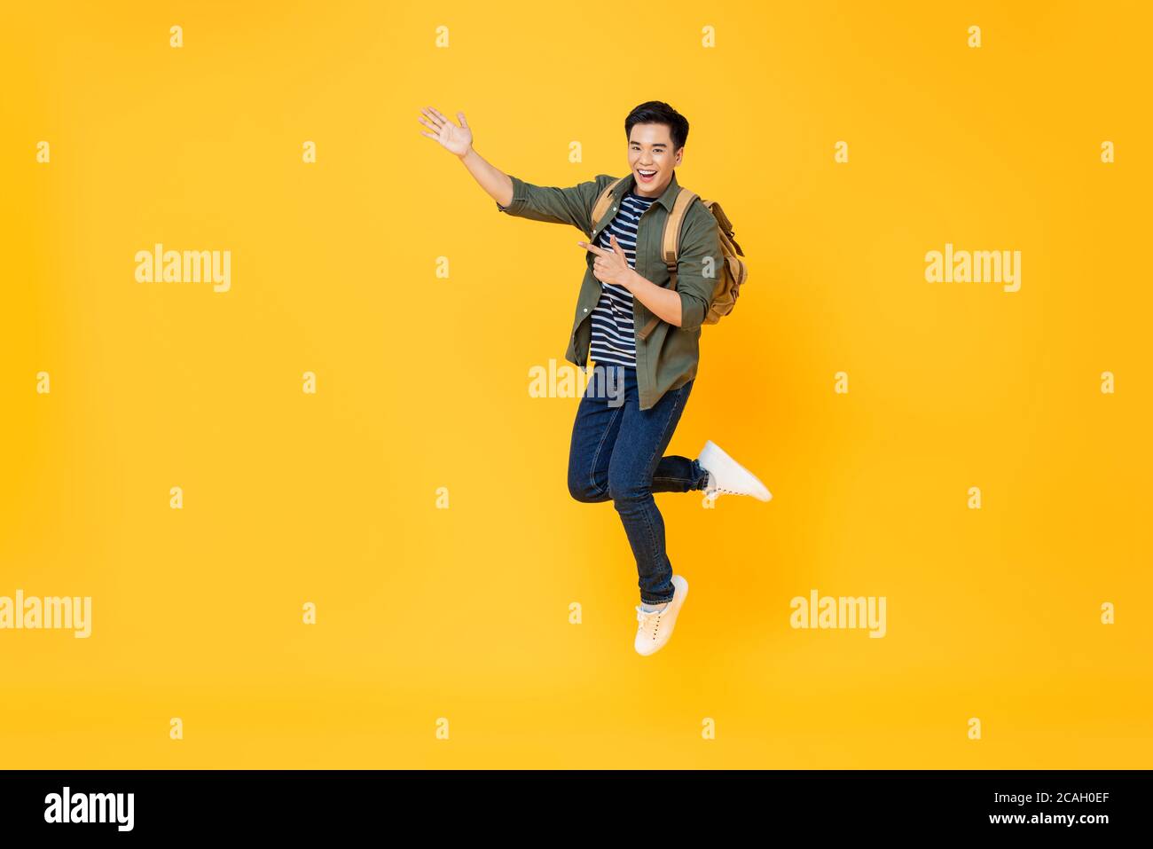 Un touriste asiatique souriant et enthousiaste avec un sac à dos qui saute et pointe les mains pour vider l'espace de côté sur un arrière-plan isolé de studio jaune Banque D'Images