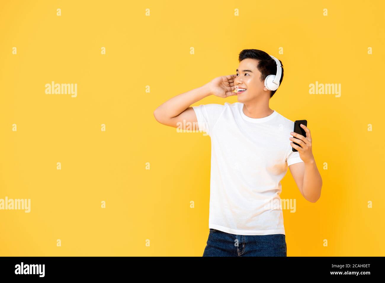 Jeune homme asiatique élégant portant un casque pour écouter de la musique depuis un smartphone sur fond jaune Banque D'Images