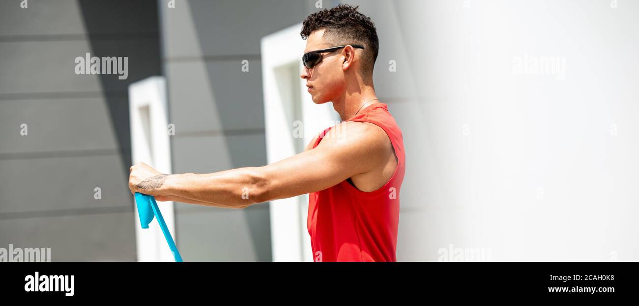 Image de bannière de beau sport homme faisant épaule avant soulever faites de l'exercice avec un bracelet de résistance à l'extérieur au soleil Banque D'Images