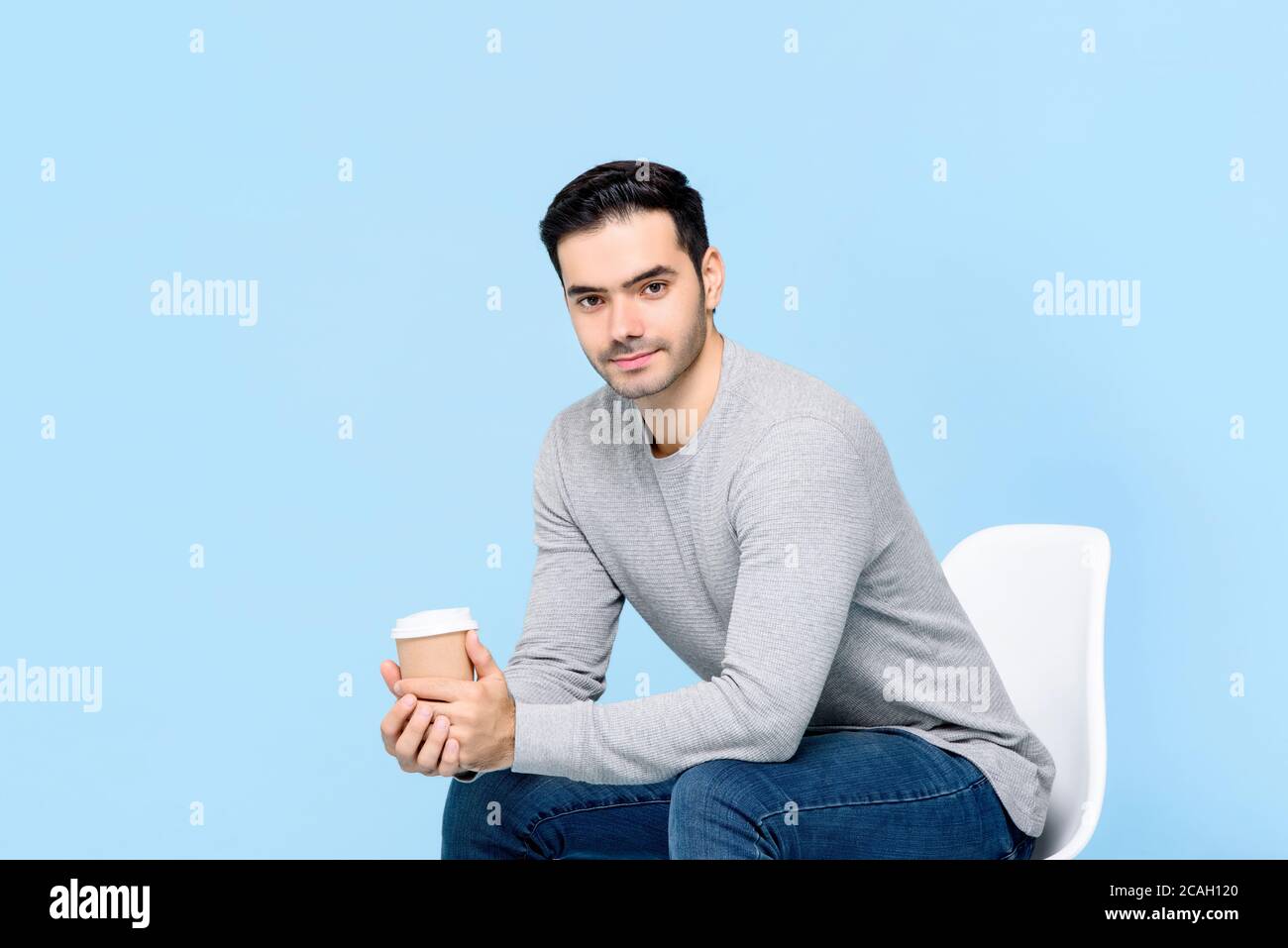 Portrait d'un jeune homme caucasien souriant regardant l'appareil photo tout en étant assis et en tenant la tasse à café isolée sur bleu clair arrière-plan du studio Banque D'Images