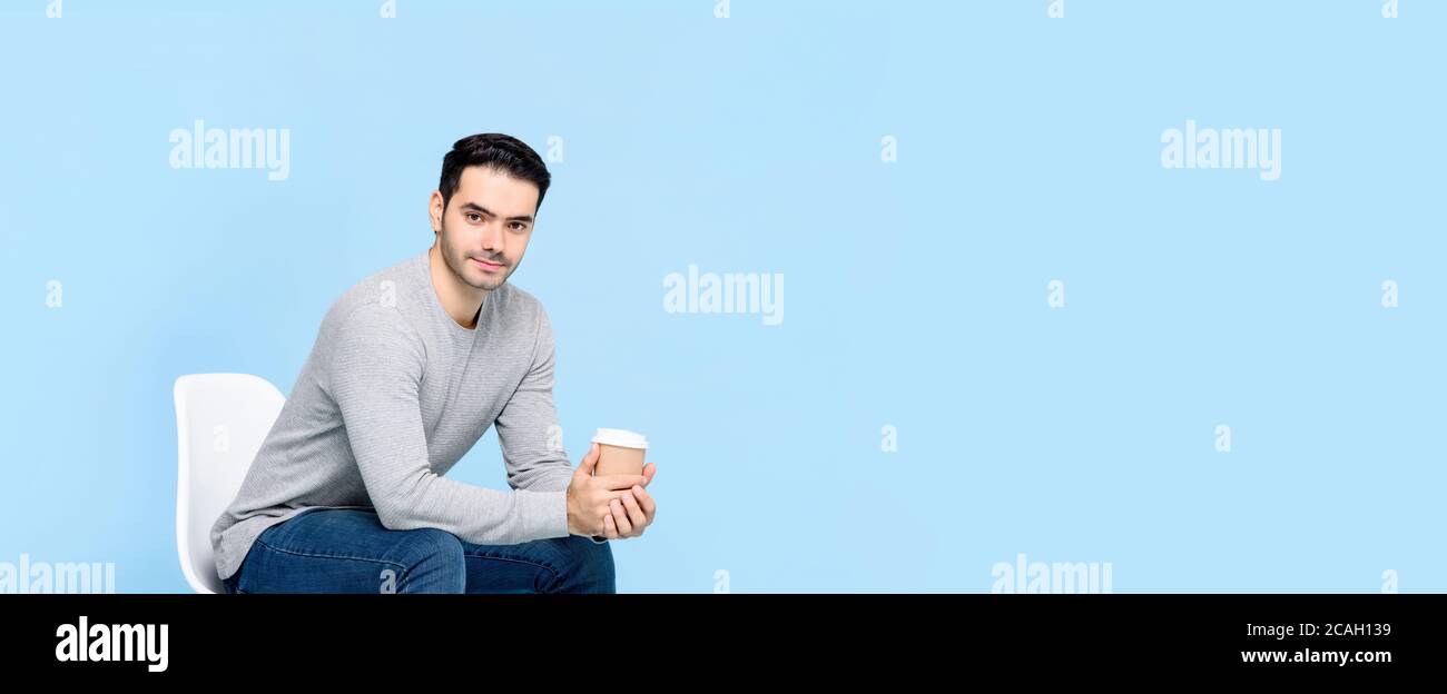 Portrait d'un jeune homme caucasien beau regardant l'appareil-photo pendant assis et tenant une tasse de café isolée sur un studio bleu clair arrière-plan de la bannière Banque D'Images