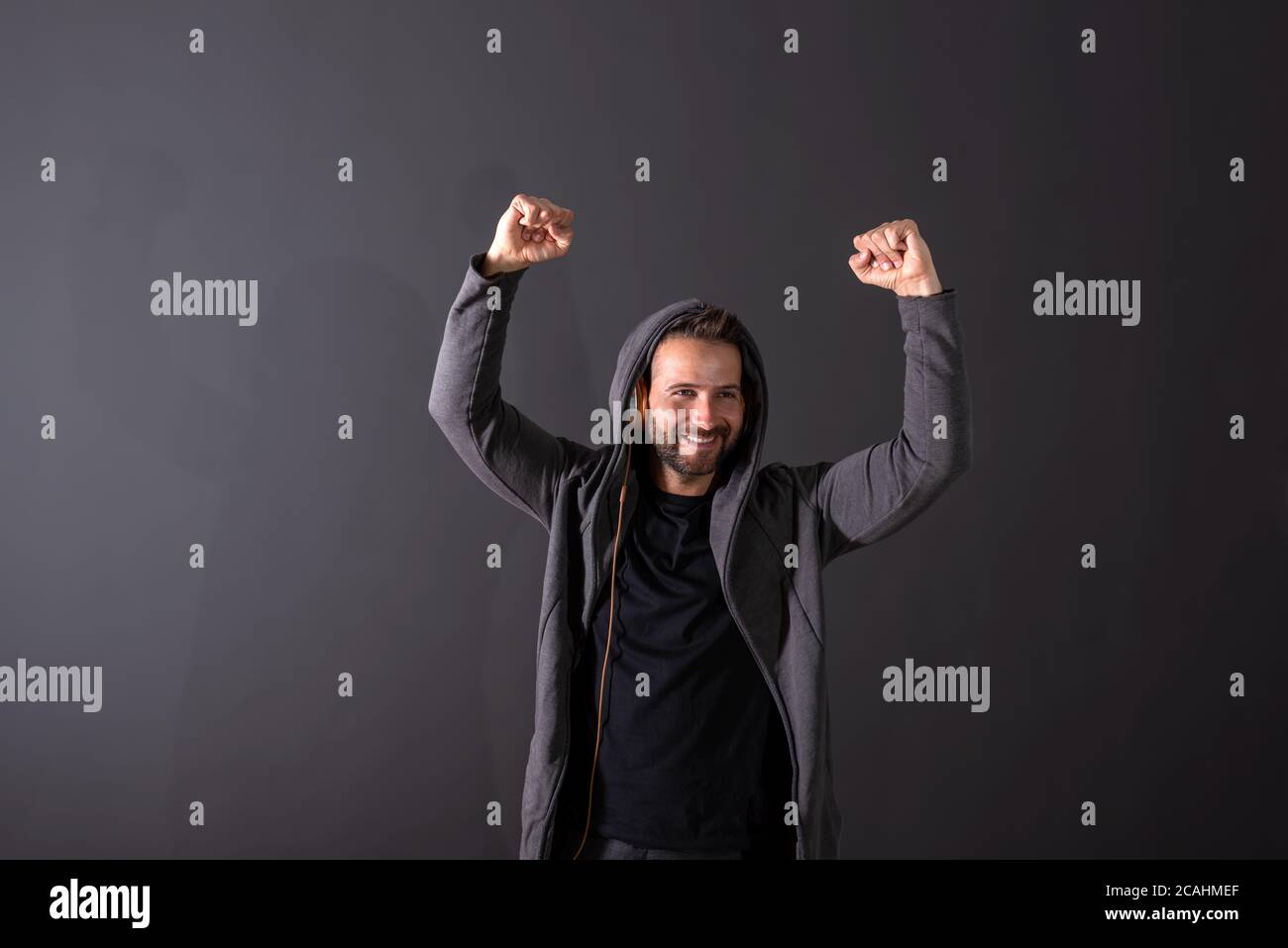 Un beau jeune homme à capuche gris qui écoute de la musique et applaudit devant un fond gris foncé dans un studio. Banque D'Images