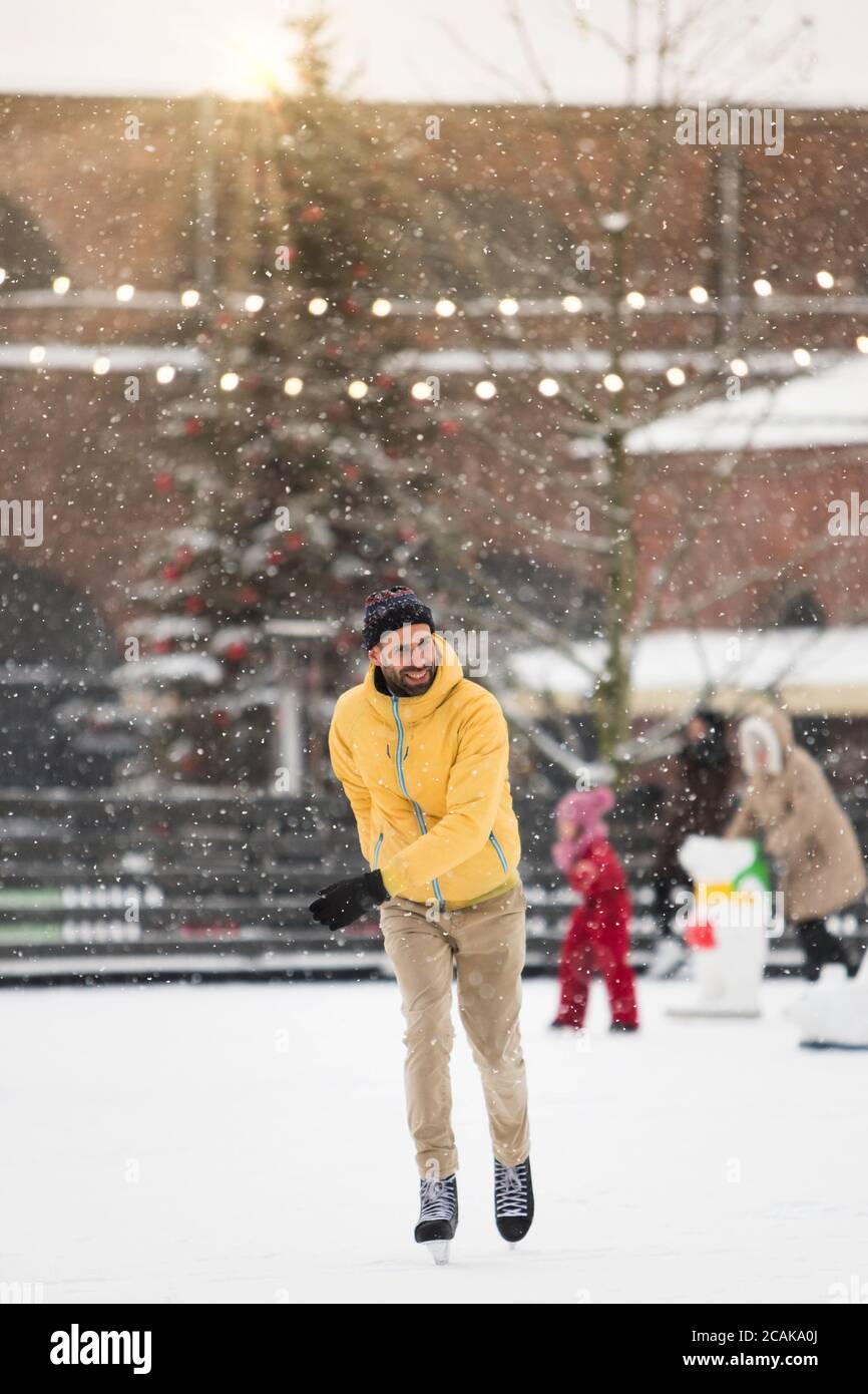 Portrait de l'homme barbu scandinave joyeux en veste jaune, pantalon beige sur patinoire, plein air en hiver enneigé, arbre de Noël en arrière-plan. Banque D'Images