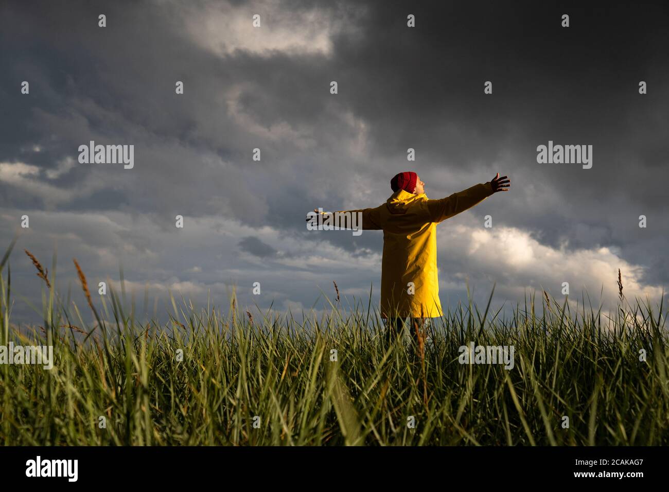 Homme en imperméable jaune et chapeau rouge avec les mains écartant largement ouvertes debout sur le terrain par temps pluvieux, regarde le ciel nuageux dramatique. Saison d'automne Banque D'Images